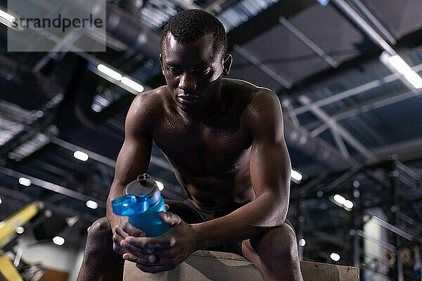 Afroamerikanischer sportlicher Mann mit Wasserflasche  der sich im Fitnessstudio entspannt