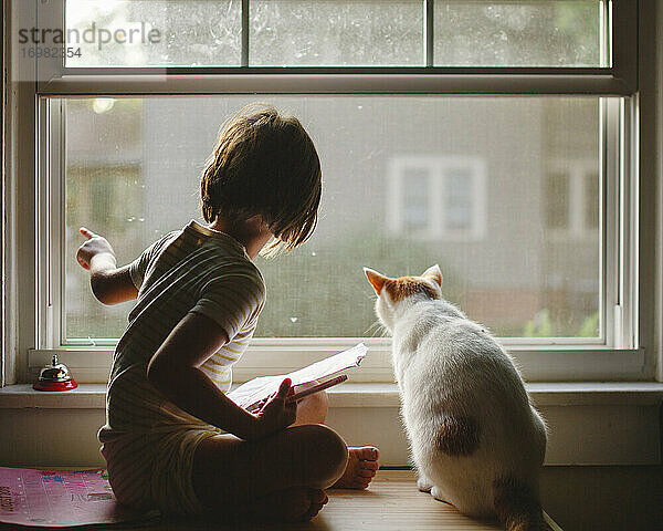 Ein kleines Mädchen sitzt auf einer Bank mit einer Katze und schaut gemeinsam aus dem Fenster