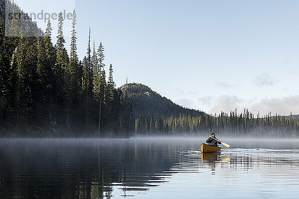 Junger Mann paddelt Kanu auf nebligem ruhigen See mit Himmel