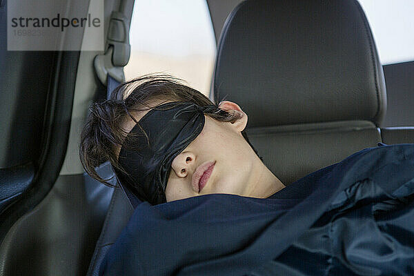 Ein Kind schläft in einem Autositz und trägt eine Augenmaske  die mit einer Decke abgedeckt ist.