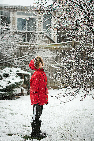 Junge in roter Winterjacke  der an einem verschneiten Tag draußen im Hof steht.