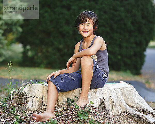 Porträt eines glücklichen barfüßigen Jungen mit goldener Haut  der auf einem Baumstumpf sitzt