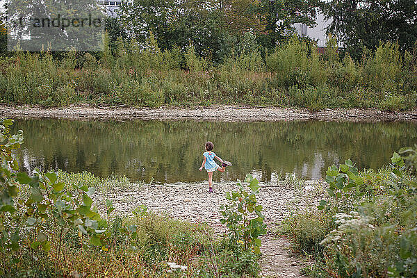 Fernansicht eines Mädchens  das am Ufer eines Flusses steht und auf einem Bein balanciert