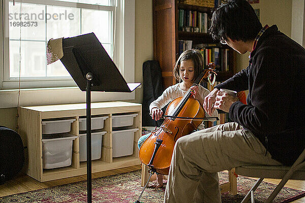 Ein Vater hilft einem kleinen Mädchen  zu Hause am Fenster Cello zu üben