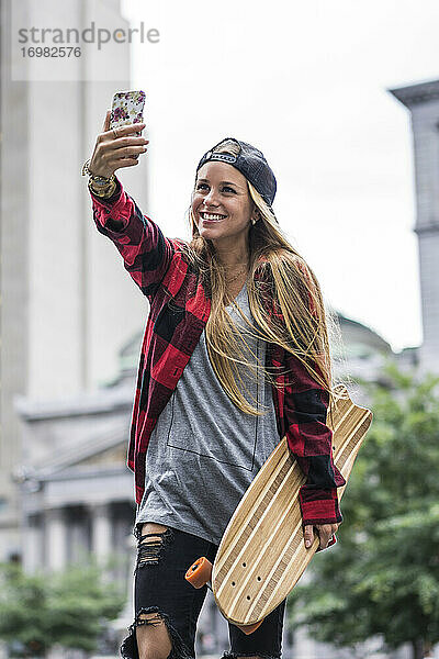 Stylische Städterin mit Skateboard macht ein Selfie in der Innenstadt