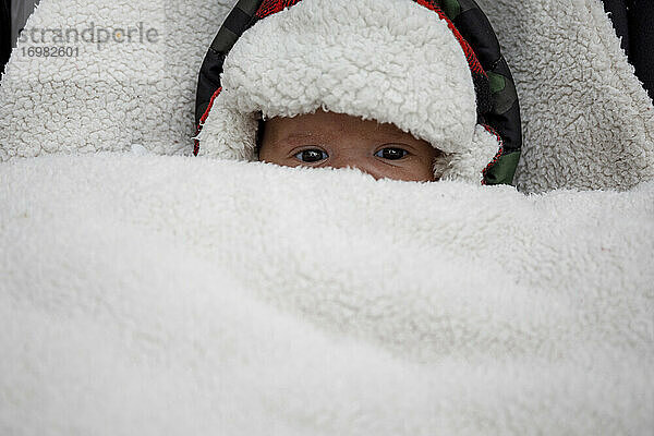Von oben von lustigen niedlichen kleinen Baby in warme Mütze mit Fell bedeckt Blick auf die Kamera  während im Kinderwagen im Winter liegen
