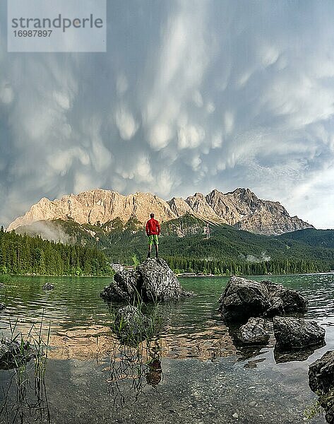 Junger Mann steht auf einem Felsen am Ufer  Eibsee vor Zugspitzmassiv mit Zugspitze  Sonnenuntergang  dramatische Mammatenwolken  Wettersteingebirge  bei Grainau  Oberbayern  Bayern  Deutschland  Europa