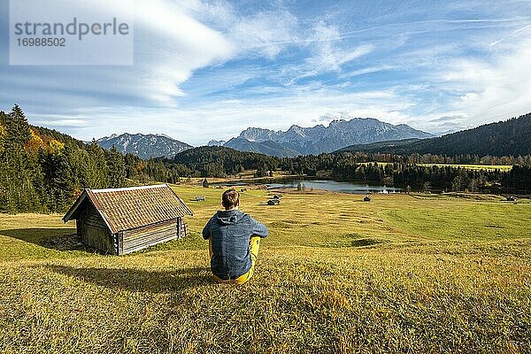 Junger Mann sitzt in der Wiese  Heustadl auf Wiese am Geroldsee  Gerold  Blick auf Karwendelgebirge  Bayern  Deutschland  Europa