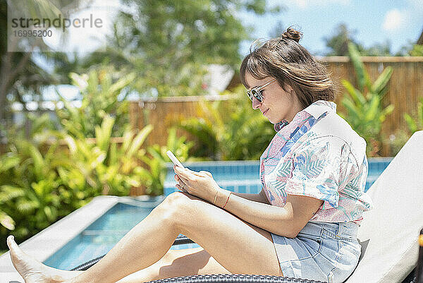 Frau mit Smartphone auf sonnigen Pool Lounge Stuhl