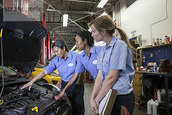 Drei Mechanikerinnen schauen in den Motorraum eines Autos in einer Werkstatt