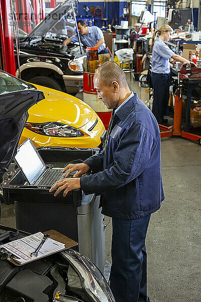 Ein pazifisch-isländischer Mechaniker tippt auf einem Laptop  während seine Kollegen im Hintergrund an Autos arbeiten