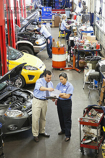 Der Besitzer einer Autowerkstatt spricht mit einer hispanischen Mechanikerin  während andere im Hintergrund arbeiten