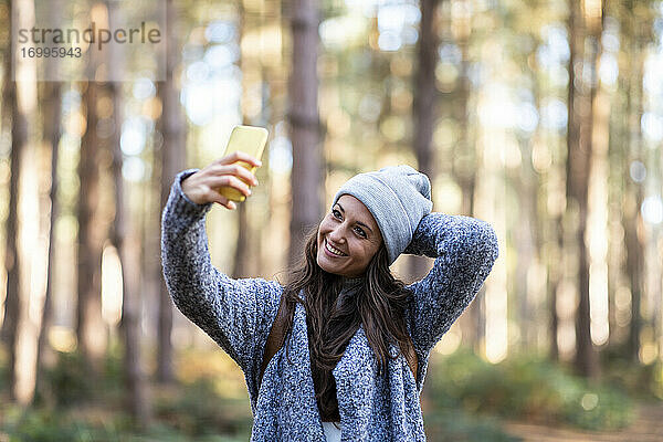 Lächelnde Frau mit Hand hinter dem Kopf  die ein Selfie macht  während sie im Wald von Cannock Chase steht