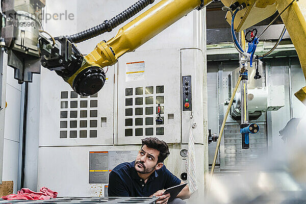 Junge männliche Arbeitskraft mit digitalem Tablet  die einen Roboterarm in einer Fabrik untersucht