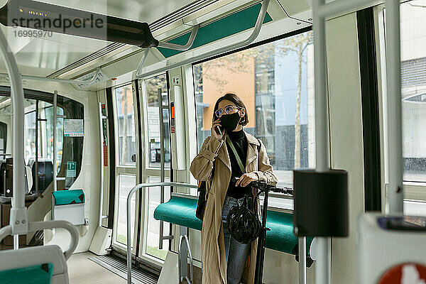 Frau mit Elektroroller und Gesichtsmaske  die in der Straßenbahn mit ihrem Handy telefoniert