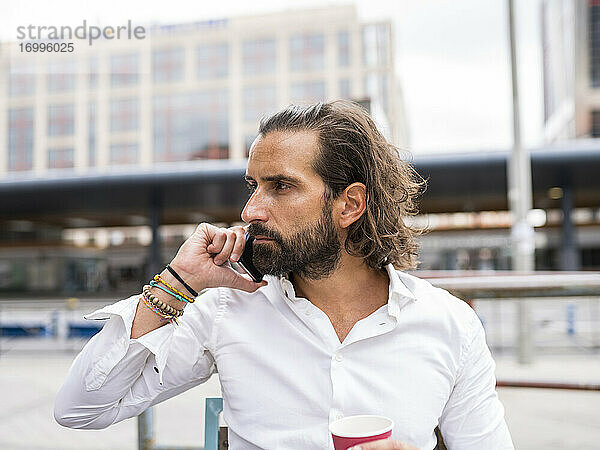 Unverfälschtes Porträt eines bärtigen Geschäftsmannes  der im Freien sitzt und mit seinem Smartphone telefoniert