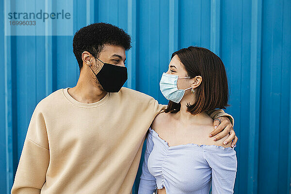Junges Paar mit Schutzmaske  das sich gegenseitig ansieht  während es an einer blauen Wand steht