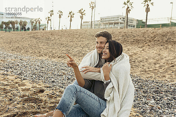 Lächelnde Freundin  die auf ihren Freund zeigt  während sie ihn am Strand umarmt