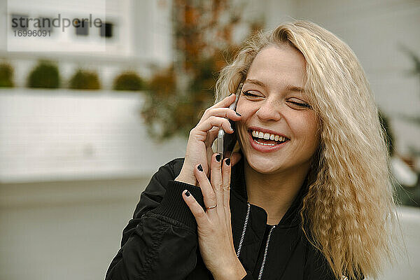 Hipster-Frau mit geschlossenen Augen  die lacht  während sie mit ihrem Smartphone im Vorgarten telefoniert