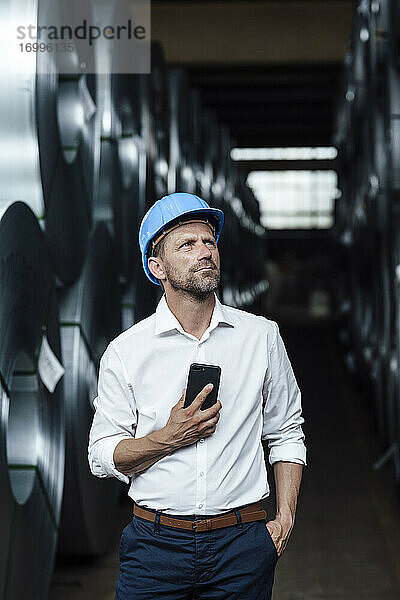 Männlicher Besitzer  der sein Smartphone hält und nach oben schaut  während er in einer Fabrik steht