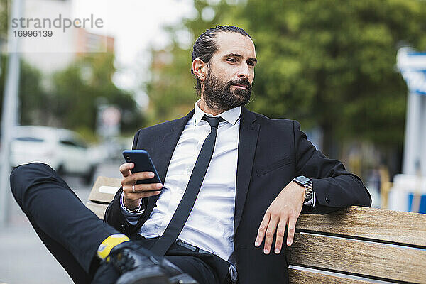 Porträt eines bärtigen Geschäftsmannes  der auf einer Bank sitzt und ein Smartphone in der Hand hält
