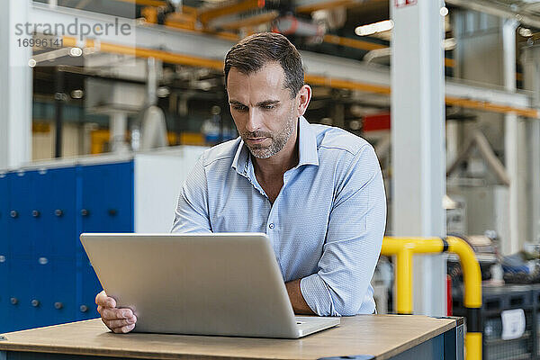 Männlicher Unternehmer  der einen Laptop benutzt und sich auf einen Schreibtisch in einer Fabrik stützt