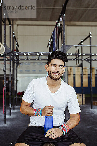 Lächelnder Mann  der eine Wasserflasche hält  während er im Fitnessstudio sitzt