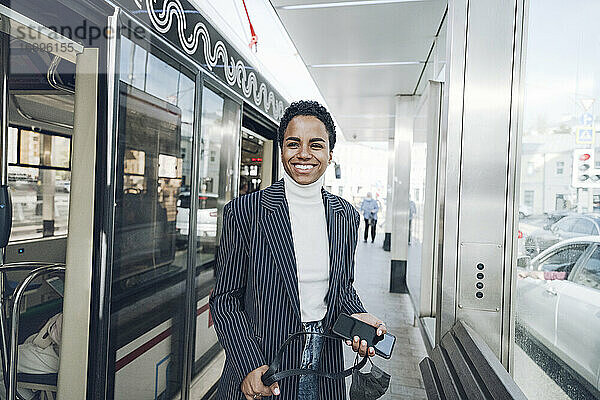 Glückliche Geschäftsfrau mit Mobiltelefon an der Bushaltestelle während des COVID-19-Ausbruchs