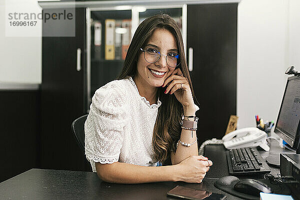 Geschäftsfrau lächelt  während sie mit dem Kopf in der Hand im Büro sitzt