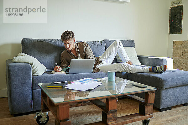 Junger männlicher Freiberufler  der im Wohnzimmer auf dem Sofa liegend mit seinem Laptop in sein Tagebuch schreibt