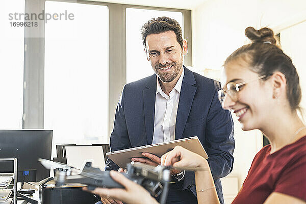 Lächelnder Geschäftsmann mit digitalem Tablet und Blick auf einen Kollegen  der einen Quadcopter hält  während er im Büro sitzt