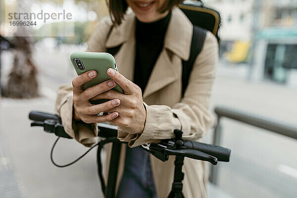 Frau  die ein Smartphone benutzt  während sie mit einem elektrischen Tretroller auf der Straße steht