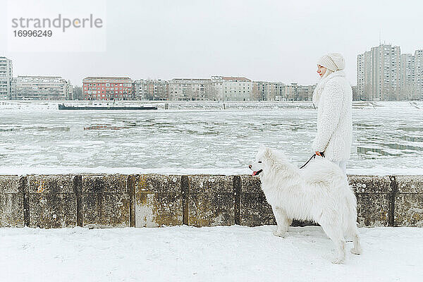 Serbien  Petrovaradin  weiß gekleidete junge Frau steht mit weißem Hund im Schnee am Flussufer