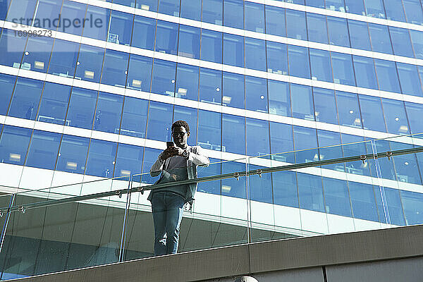 Männlicher Berufstätiger  der ein Smartphone benutzt  während er an einem Glasgeländer vor einem Bürogebäude steht