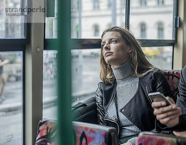 Nachdenkliche junge Geschäftsfrau  die im Bus neben einem männlichen Kollegen sitzt und sein Smartphone in der Hand hält