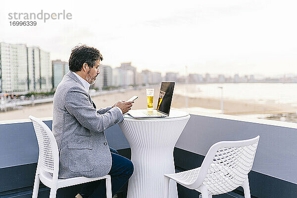 Männlicher Unternehmer  der ein Mobiltelefon benutzt  während er mit Bier und Laptop auf der Hotelterrasse sitzt