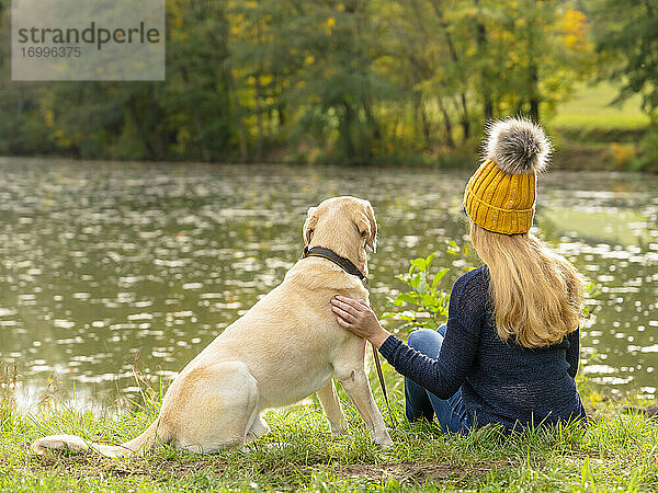 Blonde Frau sitzt mit Hund am Seeufer im Herbst