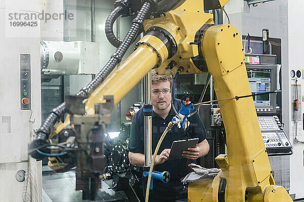 Männlicher Arbeiter  der einen Roboterarm analysiert  während er ein digitales Tablet in einer Fabrik hält