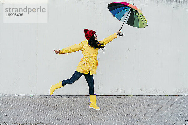 Frau mit ausgestreckten Armen  die einen bunten Regenschirm hält  während sie auf einem Fußweg an der Wand steht