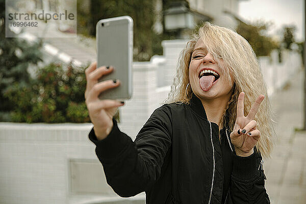 Glückliche junge Frau  die ein Selfie macht  während sie friedlich gestikuliert und die Zunge durch ihr Smartphone herausstreckt