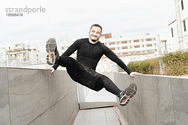 Verspielter Sportler lächelt  während er sich auf der Stützmauer eines Daches schwingt