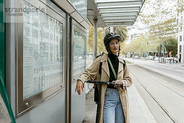 Frau mit Fahrradhelm steht mit elektrischem Roller an einer Straßenbahnhaltestelle in der Stadt