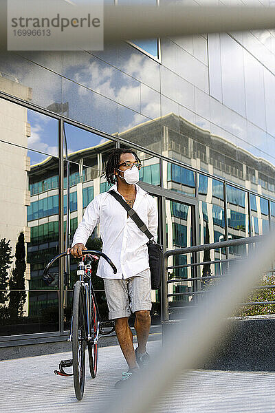 Mann mit Gesichtsmaske geht mit Fahrrad gegen modernes Gebäude während Covid-19 in der Stadt