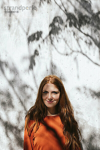 Junge Frau lächelt  während sie vor einer Baumschattenwand steht