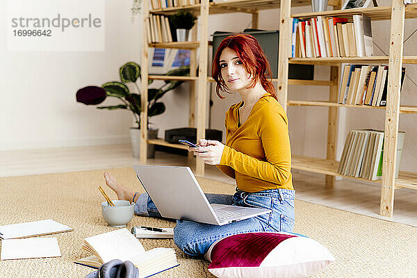Junge Frau mit Laptop auf dem Schoß  die ein Mobiltelefon benutzt  während sie zu Hause sitzt