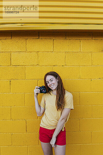 Lächelnde Frau  die eine Kamera hält und an einer gelben Wand steht