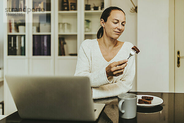 Frau isst Schokoladen-Brownie und sitzt mit Kaffee und Laptop in einer beleuchteten Küche