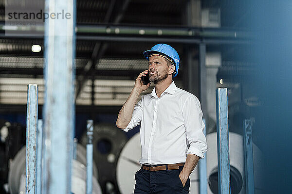 Männlicher Unternehmer  der in einer Fabrik mit der Hand in der Tasche steht und mit einem Smartphone spricht