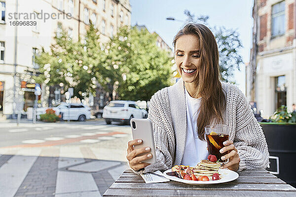 Lächelnde Frau  die ein Mobiltelefon benutzt  während sie in einem Straßencafé frühstückt