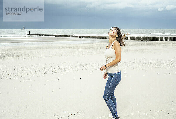 Lächelnde Frau steht mit geschlossenen Augen am Strand